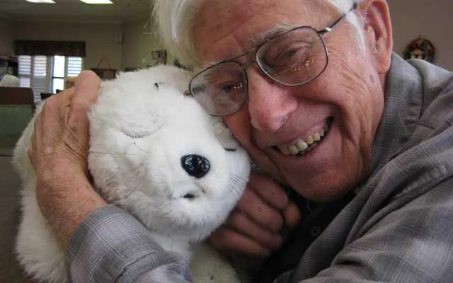 An older man smiles while holding PARO, a therapeutic robotic seal, in an elder-support setting.