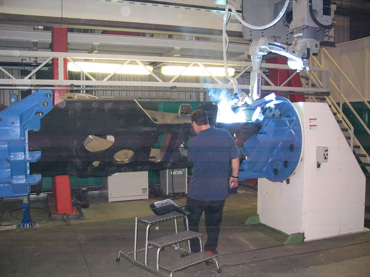 A Komatsu industrial robot produces a bright arc and smoke while welding a large metal workpiece.