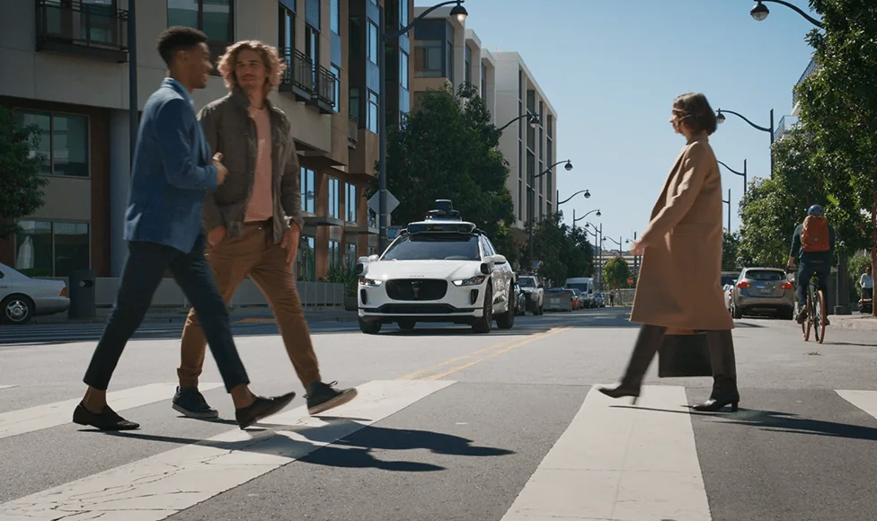 Waymo Jaguar I-PACE with its roof sensor suite approaching an urban crosswalk behind pedestrians