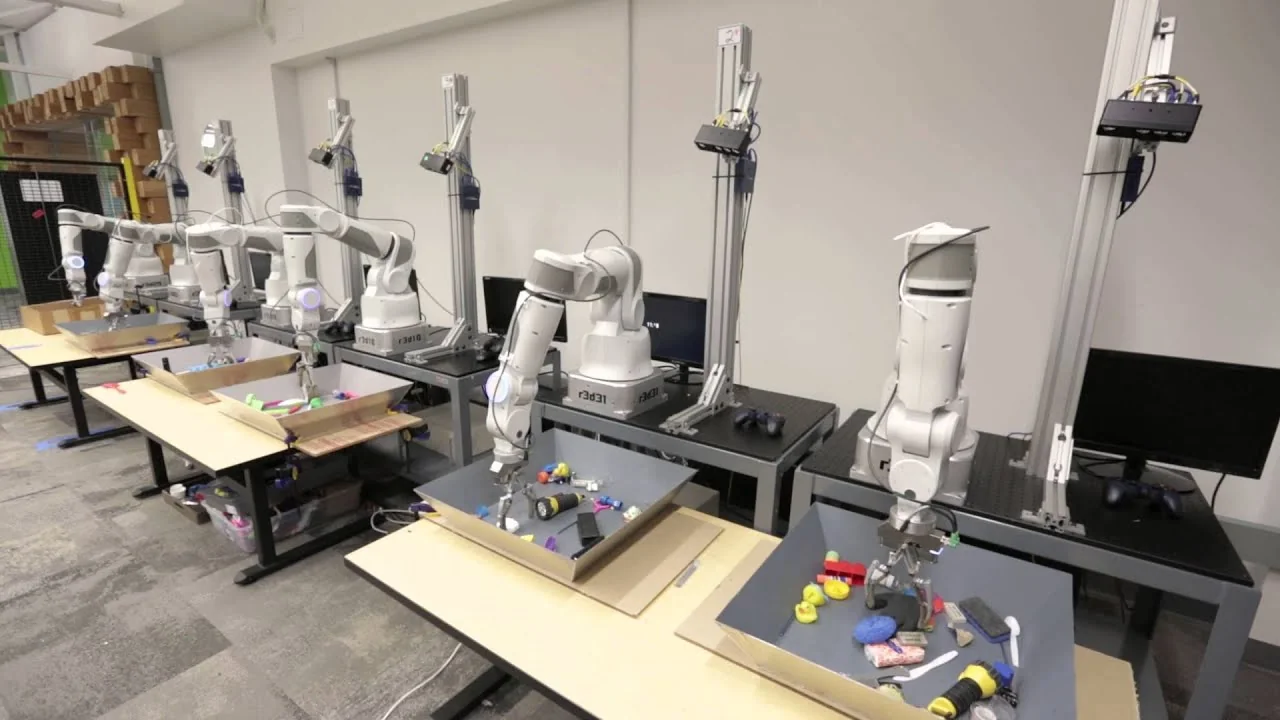 An array of robotic arms carries out parallel grasping trials over trays of household objects for the paper's large-scale data collection.